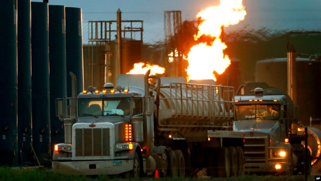 FILE - Drivers and their tanker trucks capable of hauling water and hydraulic fracturing liquid line up near a natural gas burn off flame and storage tanks in Williston, N.D., on June 9, 2014. 