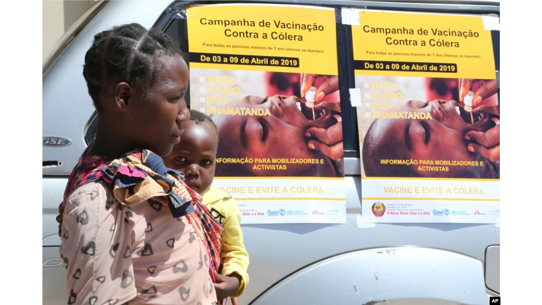 A woman and her baby walk past cholera vaccination campaign posters on the first day of the cholera vaccination program at a camp for displaced survivors of cyclone Idai in Beira, Mozambique, April, 3, 2019.