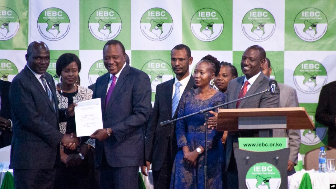 Kenyan electoral official Roselyn Akombe, center right, stands as President Uhuru Kenyatta, third from left, receives his electoral win certificate, later nullified, from Chairman of the Electoral Commission Wafula Chebukati, left, as Deputy President William Ruto, right, looks on at the results center in Nairobi, Kenya. Akombe resigned Wednesday, Oct. 18, 2017 in a statement from New York saying the rerun of the presidential election scheduled for Oct. 26 cannot be free and fair. 