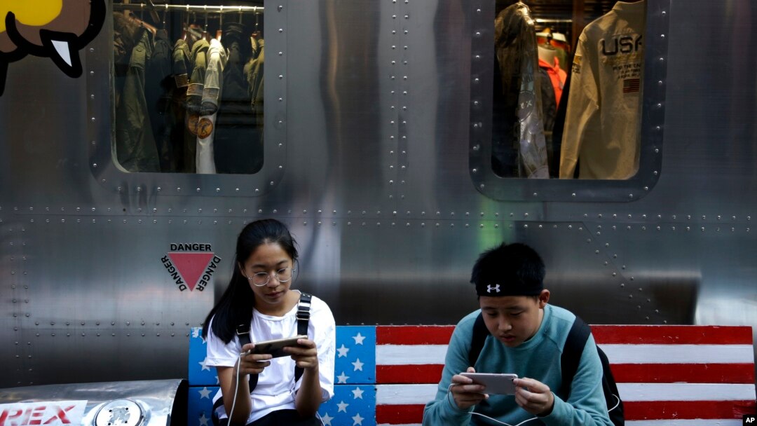 FILE - Shoppers sit on a bench with a decorated with U.S. flag browsing their smartphones outside a fashion boutique selling U.S. brand clothing at the capital city's popular shopping mall in Beijing.